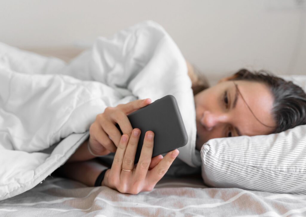 Personne allongée dans un lit regardant un téléphone portable, ce qui peut perturber le sommeil et contribuer à la fatigue